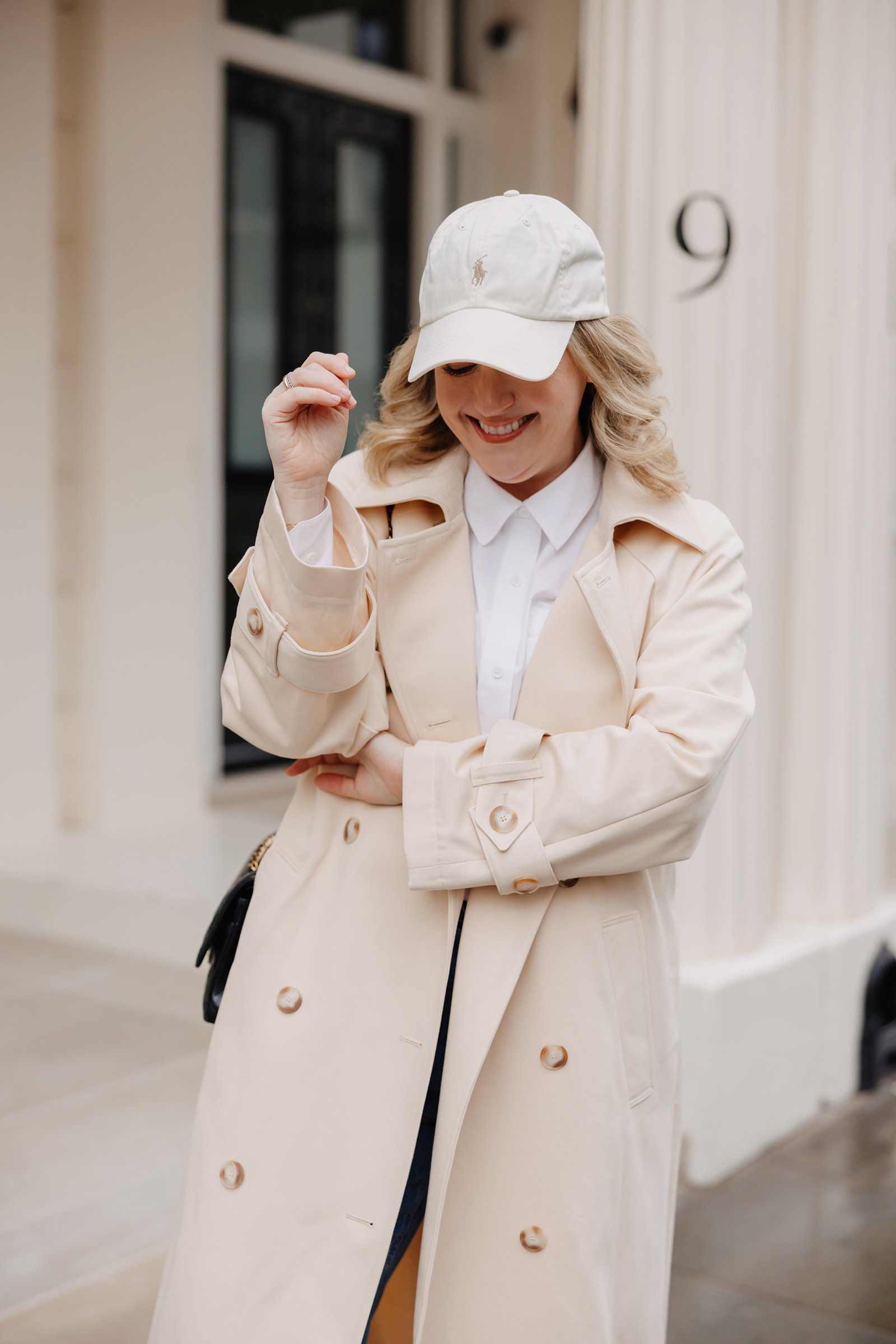 What to Wear in London Woman wearing a cream trench coat and white baseball cap in London, styled with a white button-down shirt for a classic casual London outfit.