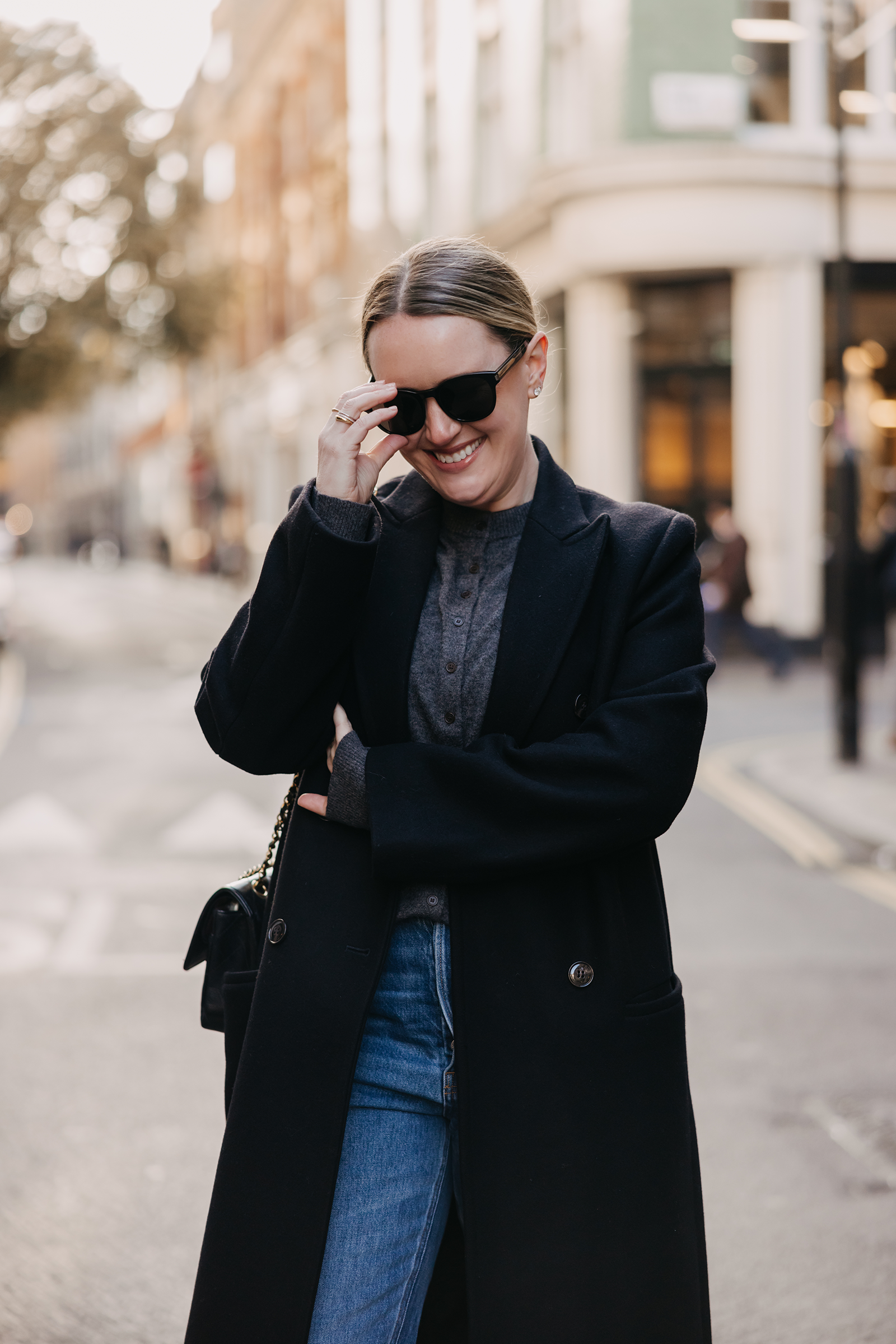 Woman wearing a black wool coat, grey sweater, straight-leg jeans, and sunglasses on a London street, showcasing a chic winter outfit for a London packing list