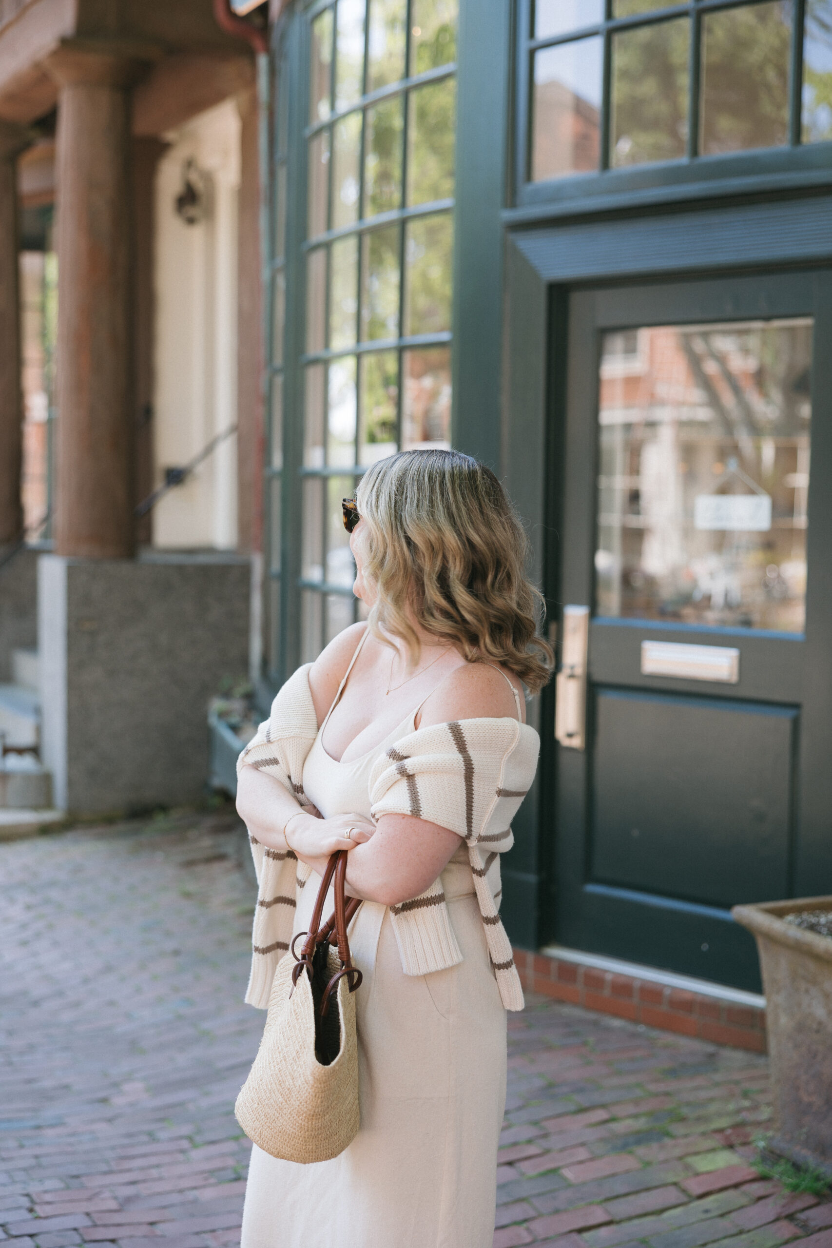 Best Sweaters for Spring and Summer Woman wearing a cream knit tank dress with a striped cardigan draped over her shoulders and carrying a woven straw tote bag on a charming brick-lined street, demonstrating effortless spring sweater styling.