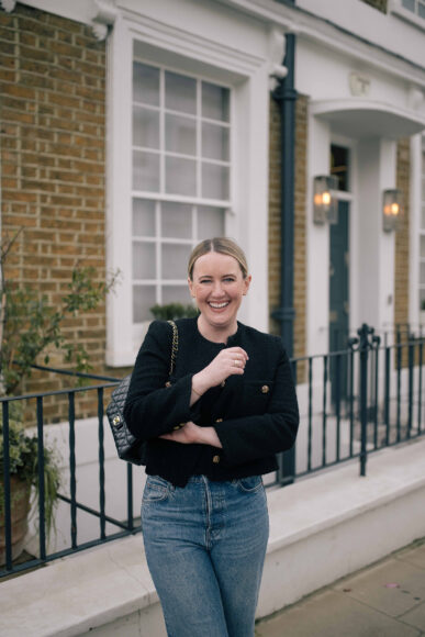 Woman wearing a black cropped tweed jacket, blue jeans, and black quilted shoulder bag smiling on a London street