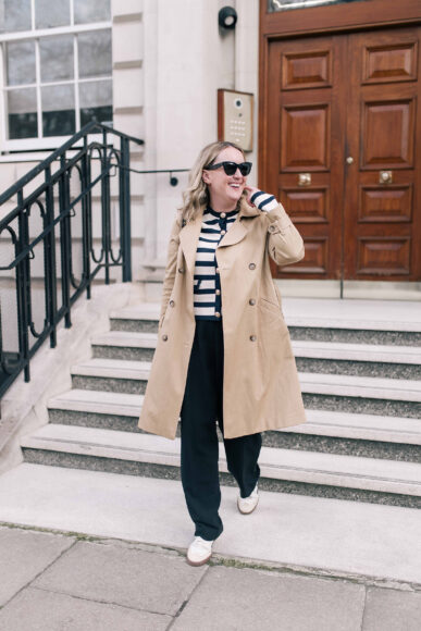 Woman wearing a beige trench coat, navy striped sweater, wide-leg trousers, and white sneakers walking confidently down city steps