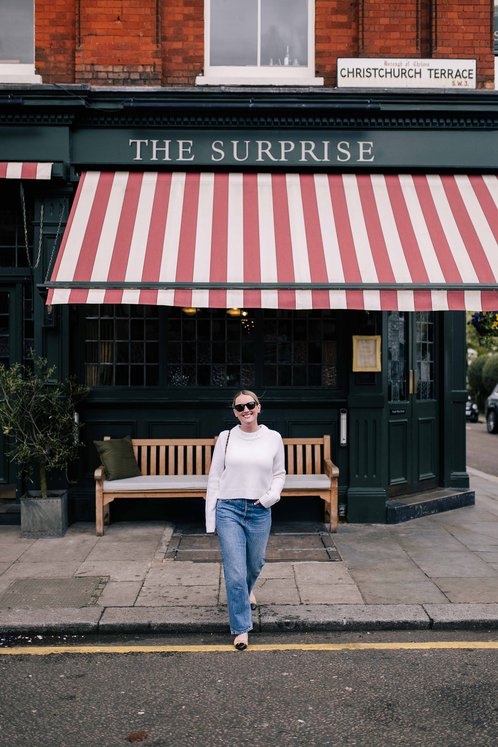 Woman wearing a white sweater, blue jeans, and cap-toe flats walking in front of The Surprise pub in London