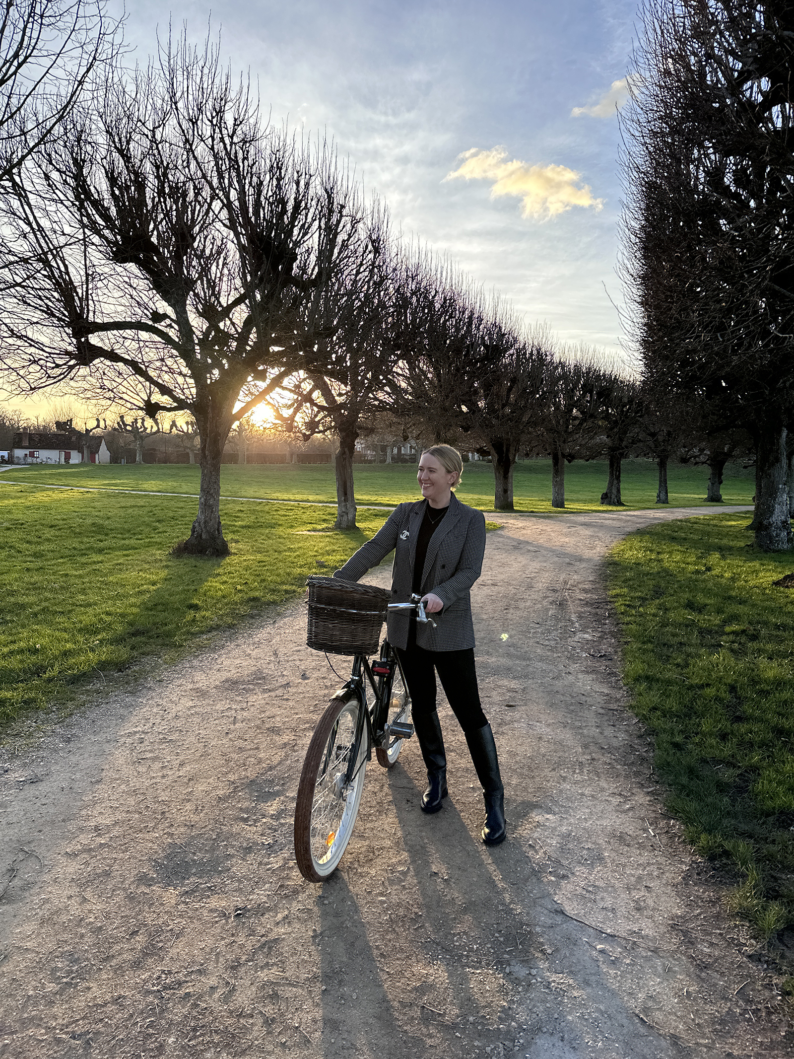 Woman wearing a plaid blazer, black turtleneck, and knee-high boots standing with a bicycle at sunset on a tree-lined path in the countryside